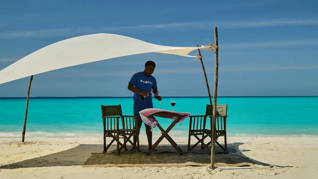 A waiter pouring wine at a table for two laid under a white sail shade on the beach