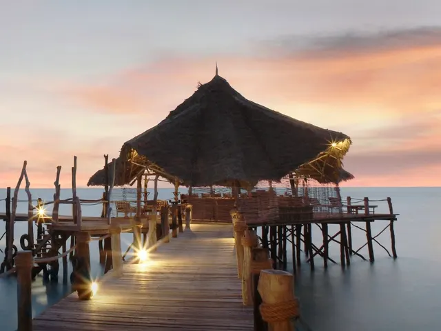 A lantern-lit jetty running out to a thatched over-water pavilion at sunset