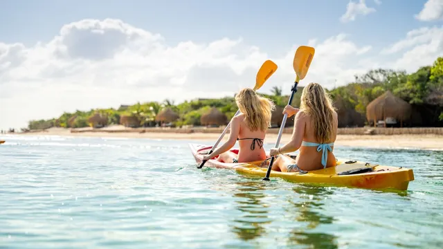 Two paddlers in a kayak on a calm lagoon with thatched lodge bandas along the beach behind them